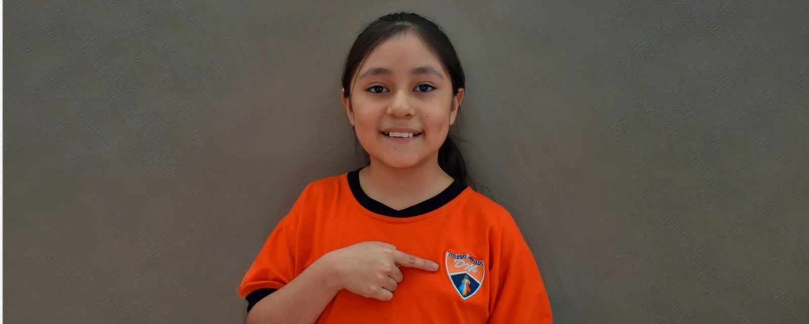 Una niña señala el escudo de su polo escolar naranja.
