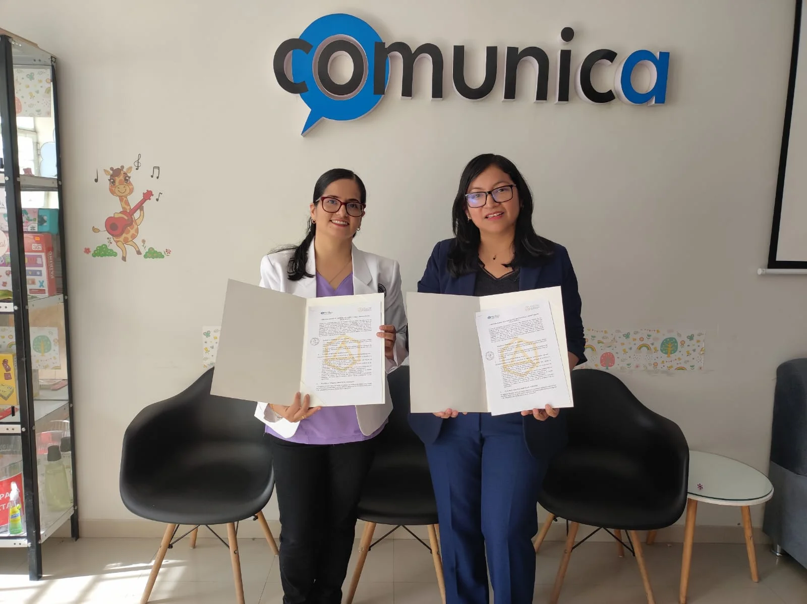 Dos mujeres sostienen documentos de convenio frente al logotipo de COMUNICA.